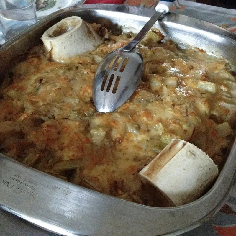 Fuente metálica con cardos cocidos gratinados y un hueso de tuétano, con una espumadera