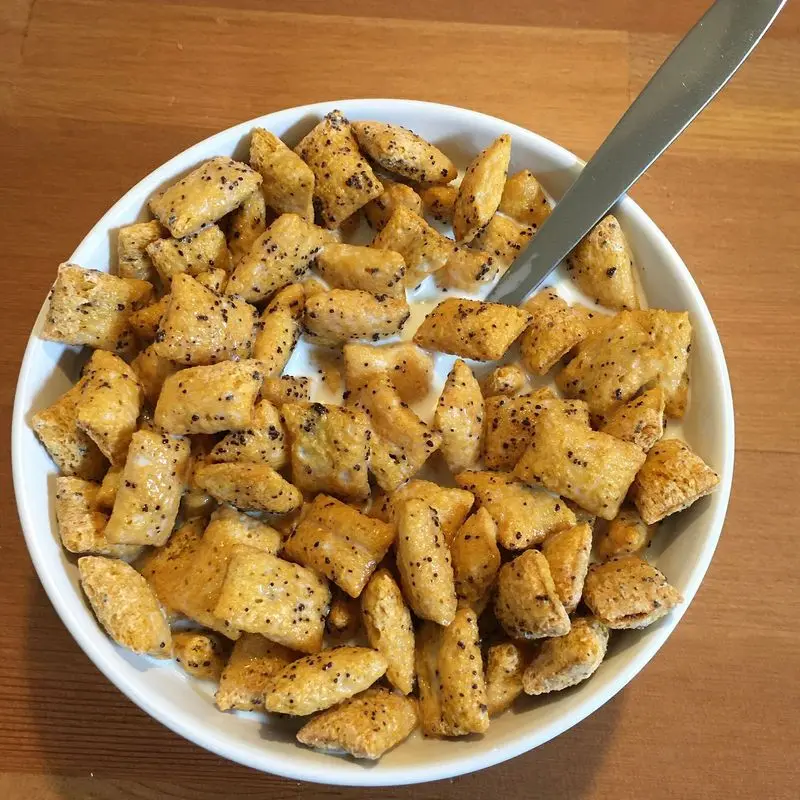 Cuenco blanco de cereales dorados en forma de almohadilla con cuchara sobre mesa de madera