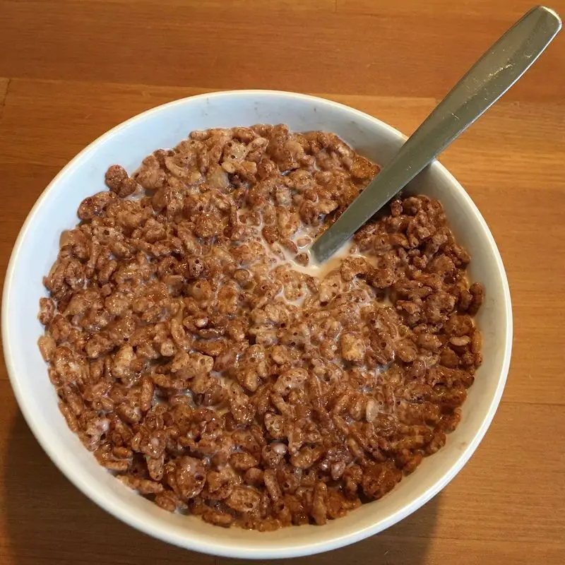 Cuenco blanco de cereales de cacao con leche y una cuchara sobre mesa de madera