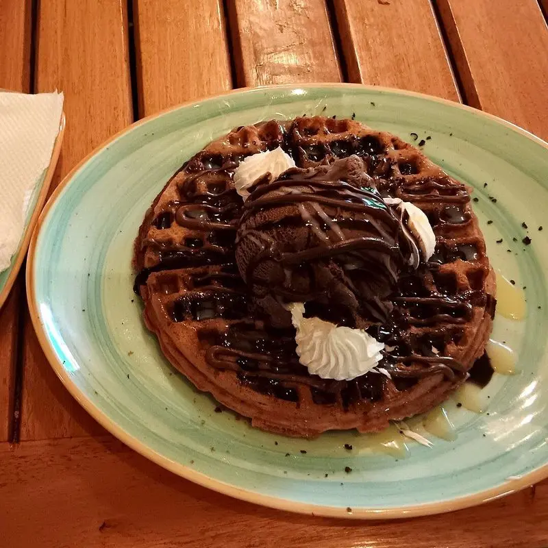 Gofre cubierto de salsa de chocolate y nata en un plato verde sobre mesa de madera