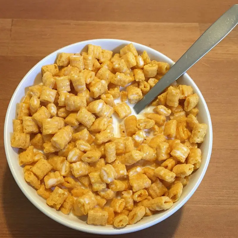Cuenco blanco de cereales dorados en forma de cuadraditos con leche y una cuchara