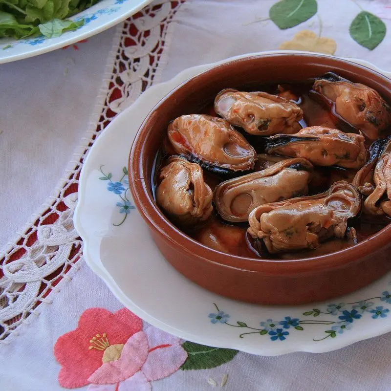Cazuela de barro con mejillones en escabeche sobre un plato y un mantel bordado