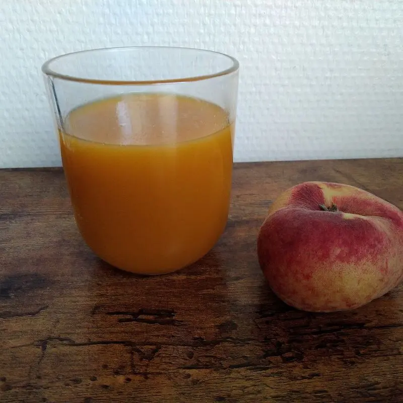 Vaso de néctar de melocotón junto a un melocotón plano sobre una mesa de madera