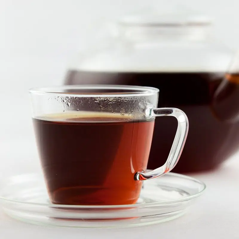 Taza de cristal con infusión de rooibos rojiza y tetera de cristal detrás