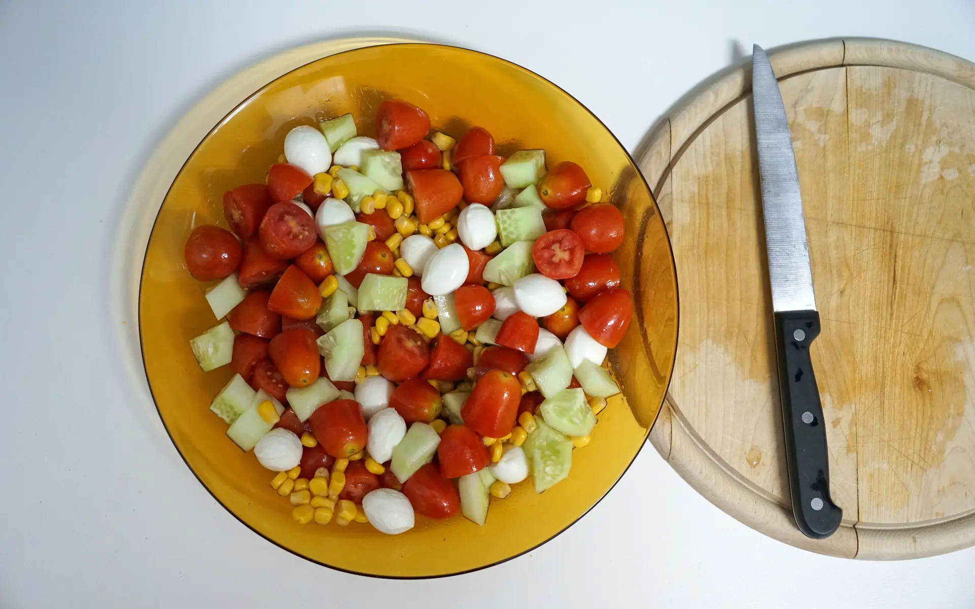 Bol de ensalada con tomate cherry, mozzarella, pepino y maíz junto a una tabla