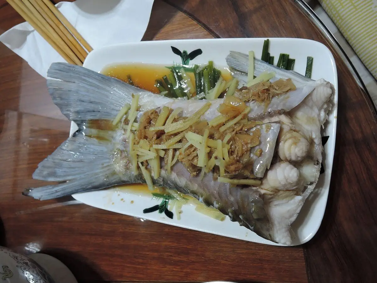Pescado entero al vapor con tiras de jengibre y salsa de soja en un plato blanco rectangular