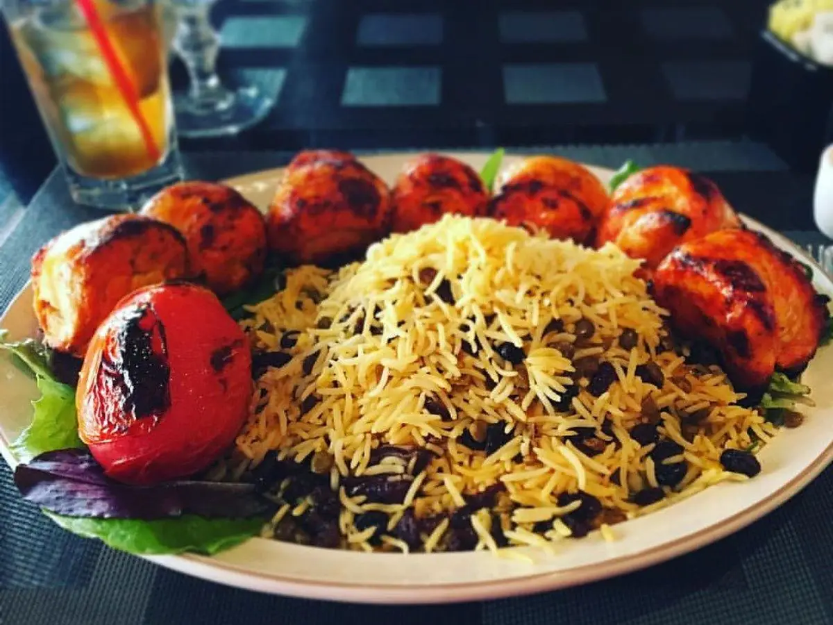 Plato de arroz persa con lentejas y pasas junto a brochetas de carne y tomates asados