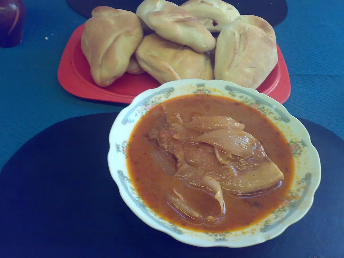 Cuenco de adobo arequipeño de cerdo en caldo rojizo con panes de tres puntas al fondo