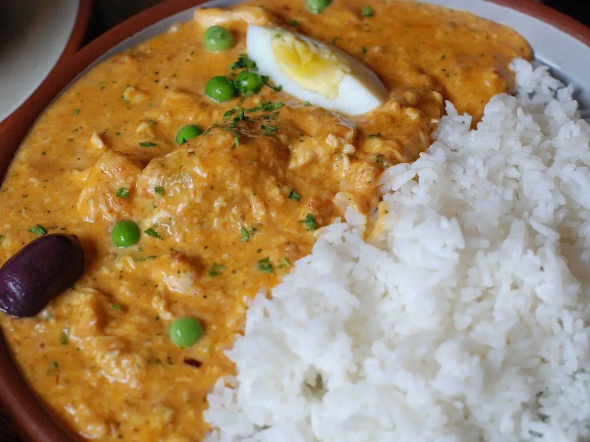 Guiso de pollo en salsa cremosa amarilla con huevo cocido, guisantes y aceituna junto a arroz blanco