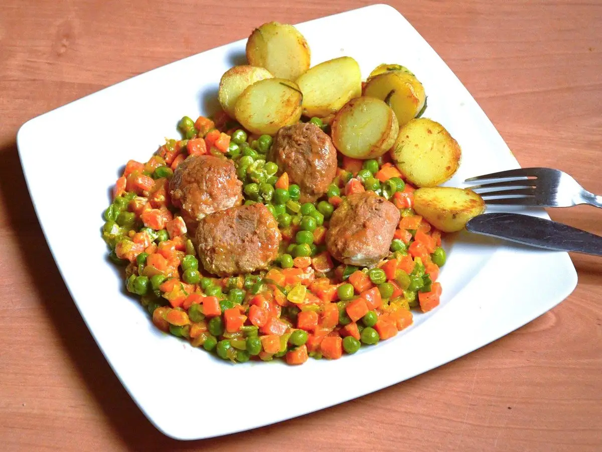 Plato blanco con albóndigas, patatas pequeñas y guisantes con zanahoria en dados