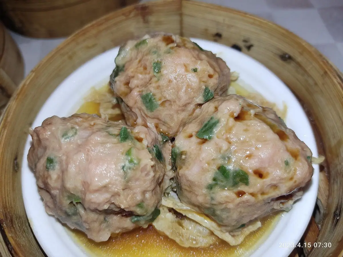 Tres albóndigas de carne al vapor con cebolleta picada sobre un plato blanco en una cesta de bambú