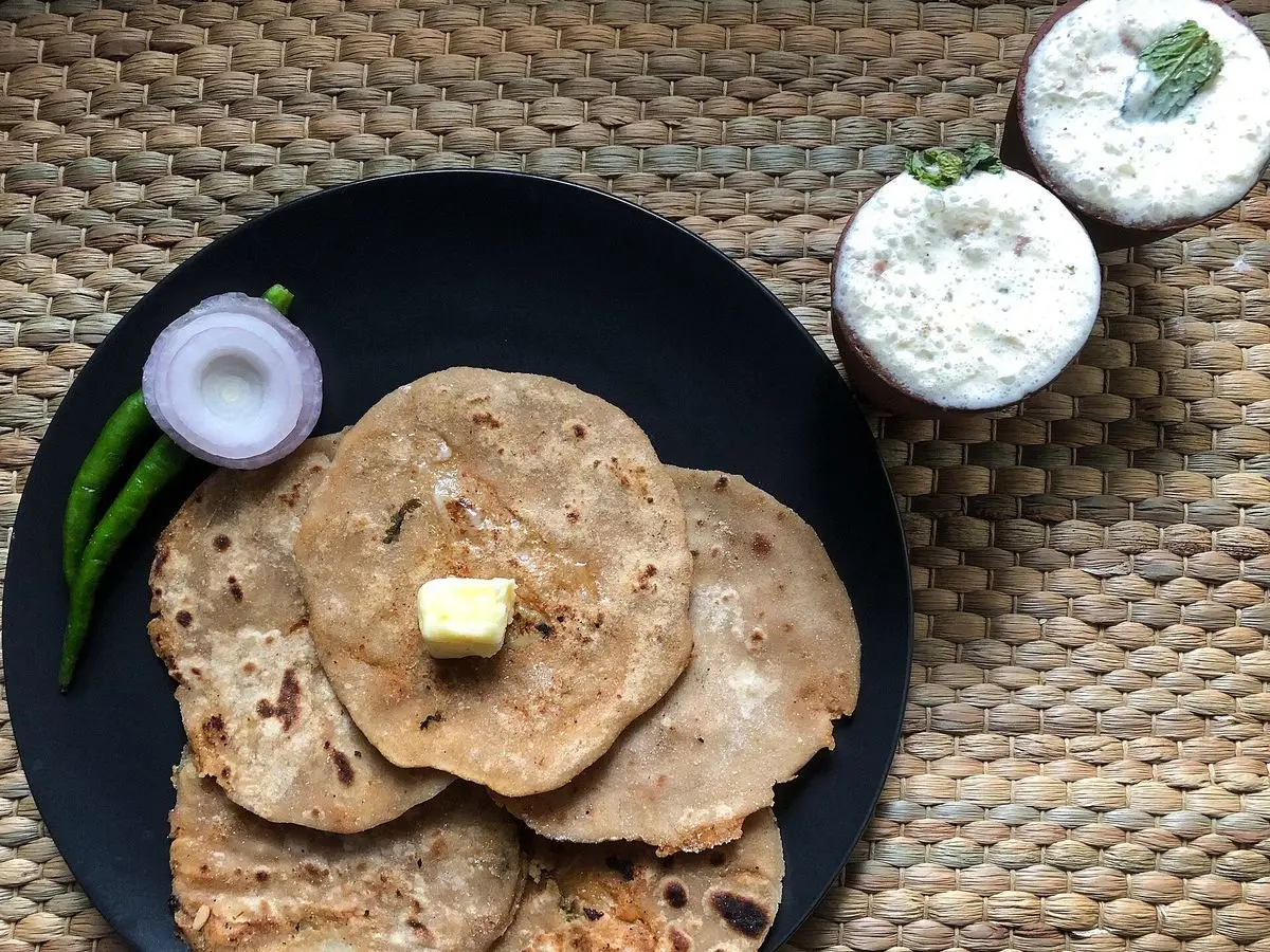 Panes indios parathas con un trozo de mantequilla, cuenco de yogur, guindilla verde y cebolla sobre un plato negro