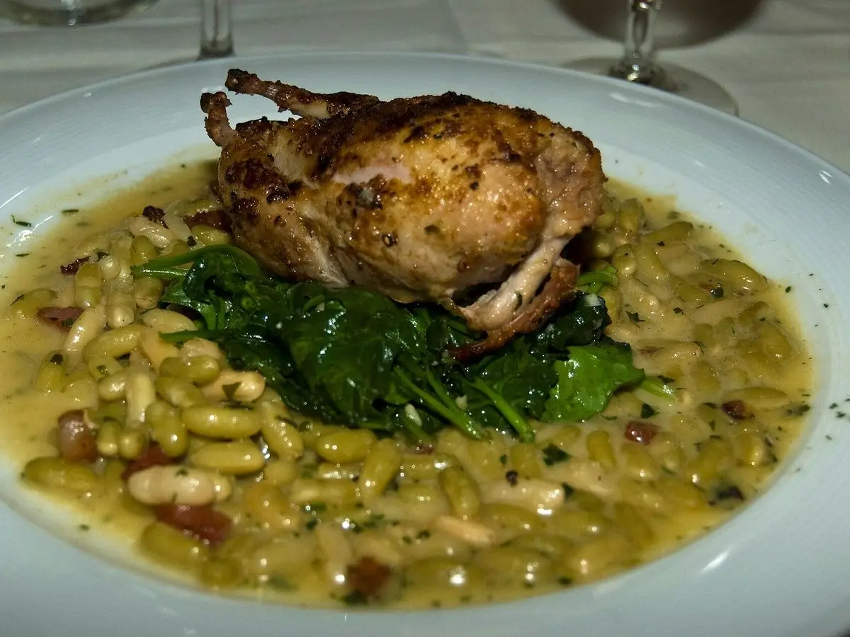 Codorniz asada sobre una cama de alubias pequeñas y espinacas en un plato blanco hondo