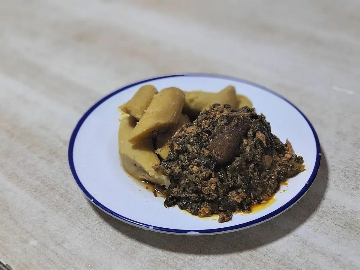 Plato blanco con plátano cocido y guiso verde oscuro de hojas, ampesi con kontomire