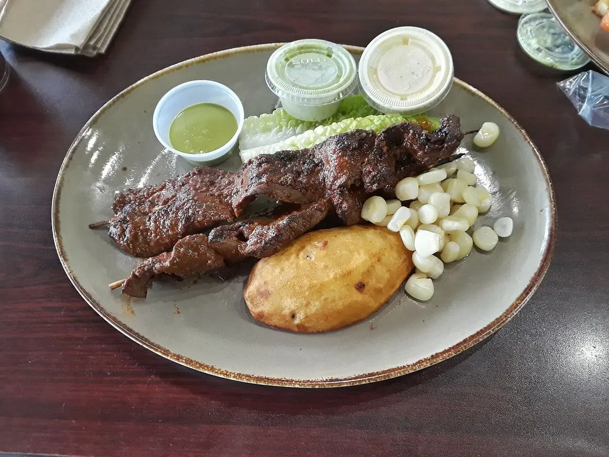 Plato con carne de anticucho a la parrilla, una patata, granos de maíz grandes y salsas verdes