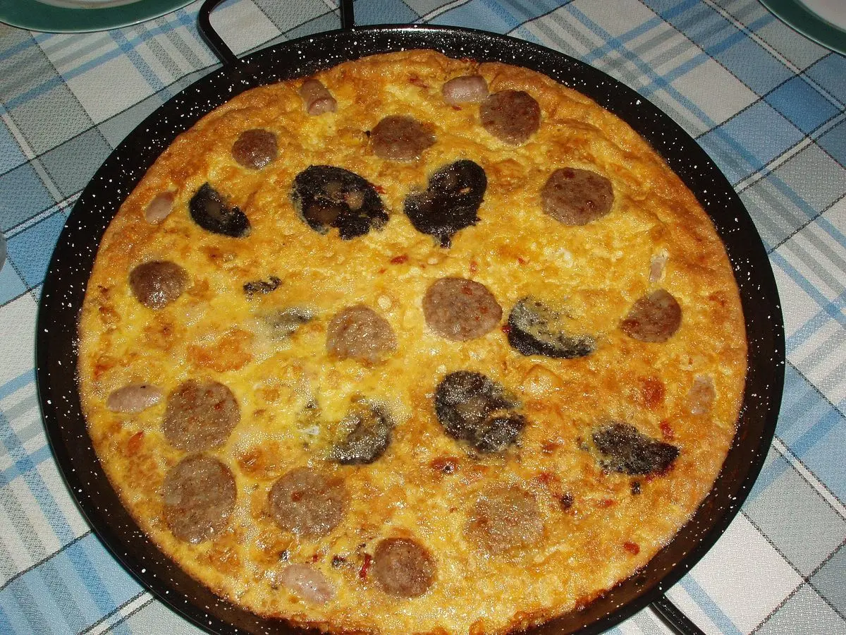 Paellera con arroz dorado con costra y rodajas de embutido y morcilla sobre un mantel de cuadros