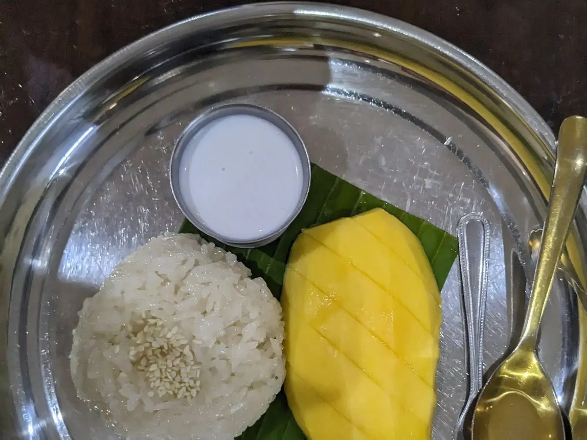 Arroz glutinoso con mango maduro, leche de coco en un cuenquito y sésamo sobre hoja de plátano