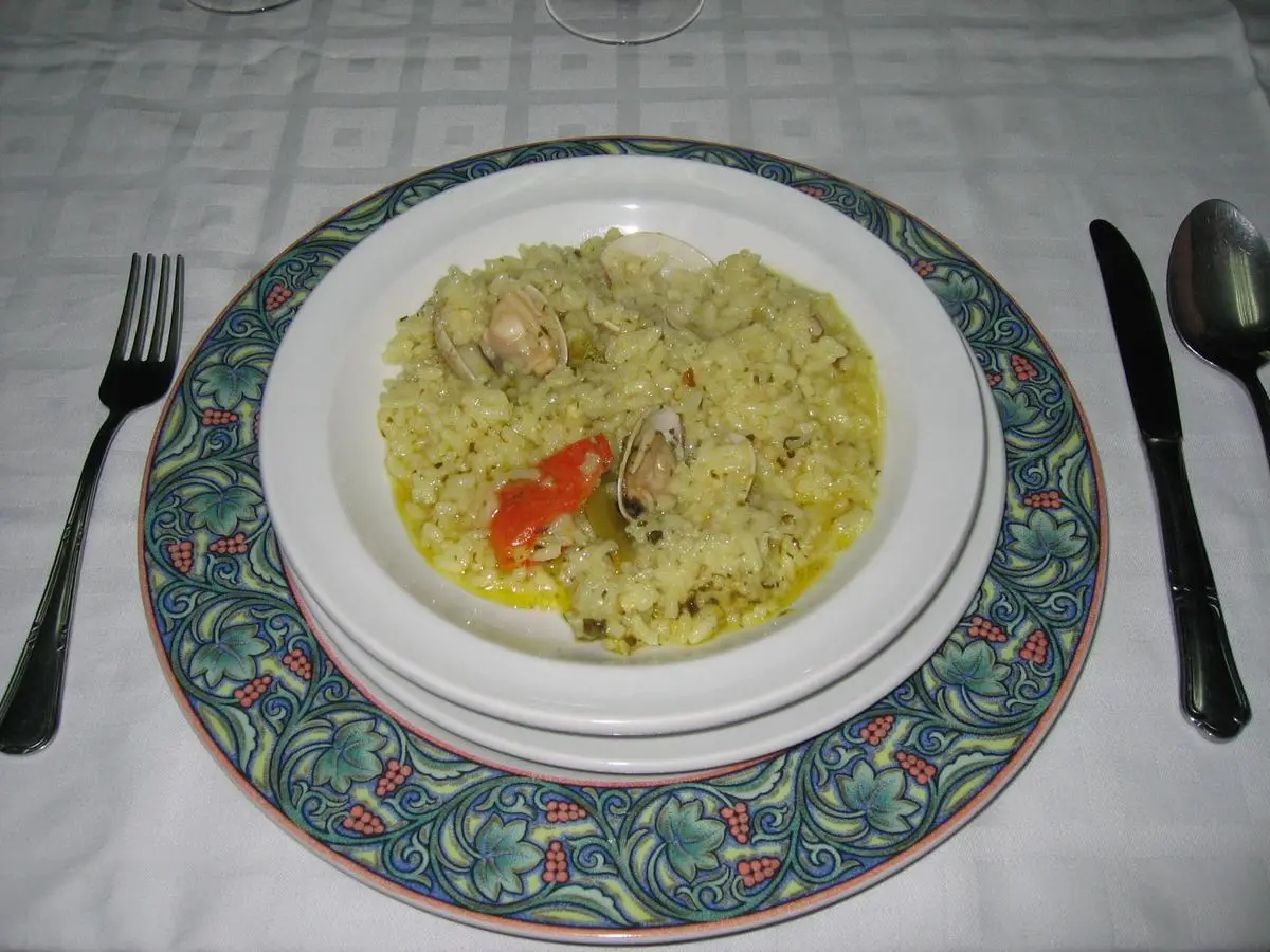 Plato de arroz caldoso verdoso con almejas y tomate sobre mantel blanco con cubiertos