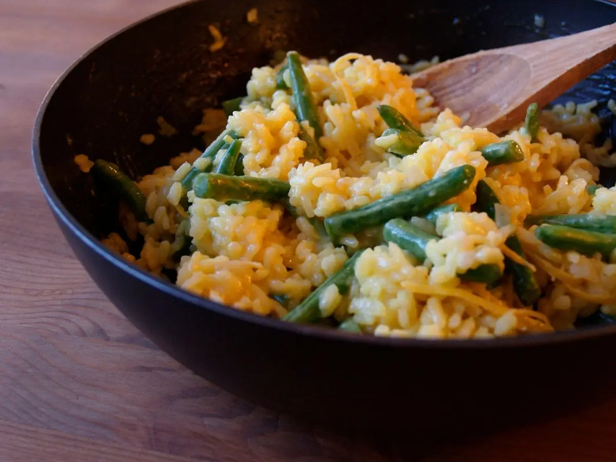 Cuenco oscuro de arroz cremoso anaranjado con judías verdes y cuchara de madera