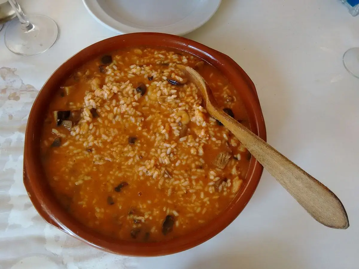 Cazuela de barro con arroz meloso con carne y setas y una cuchara de madera sobre mantel blanco