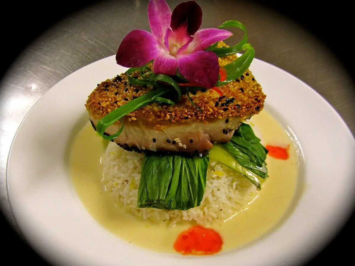 Atún sellado con costra de sésamo sobre arroz y verdura verde con salsa cremosa y una flor comestible
