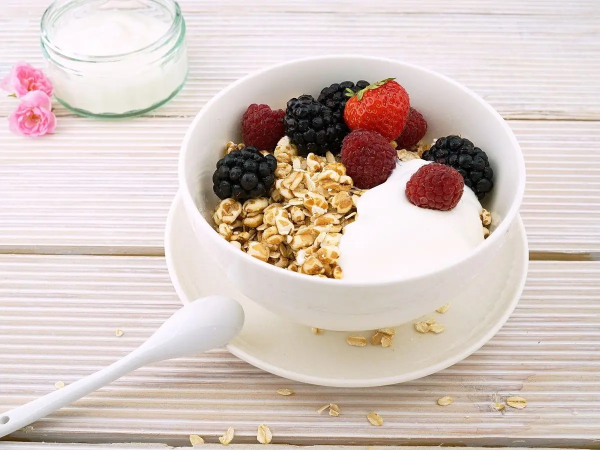 Cuenco blanco de muesli con yogur y frutos rojos, frambuesas y moras, junto a un vaso de yogur