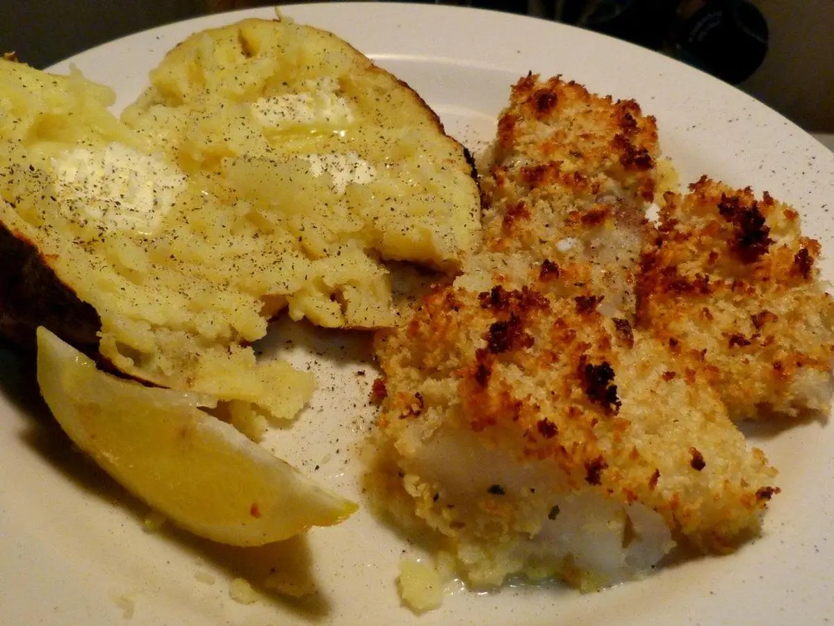 Pescado blanco al horno con costra de pan rallado dorada, junto a patata asada y limón en un plato blanco