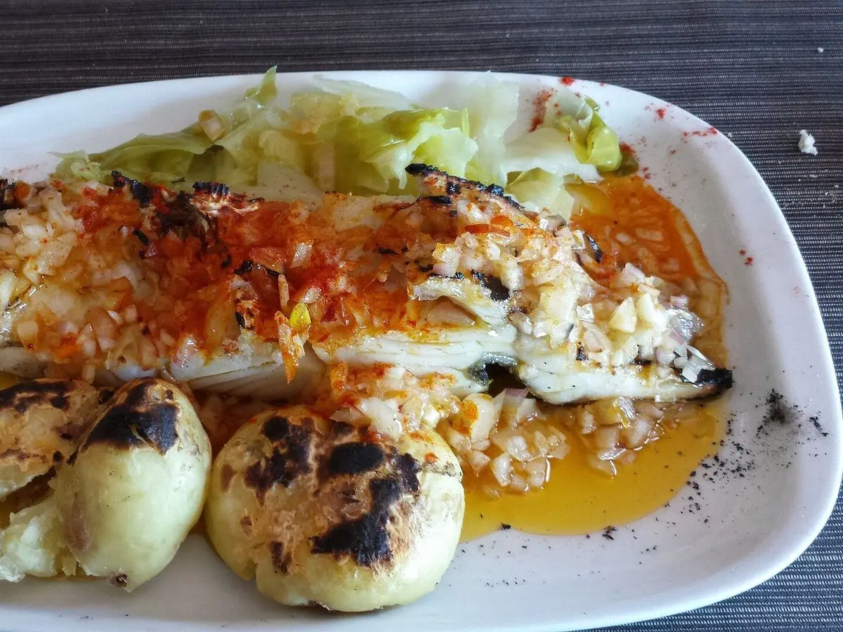 Lomo de bacalao asado con pimentón, patatas chafadas doradas y hojas verdes en una fuente blanca