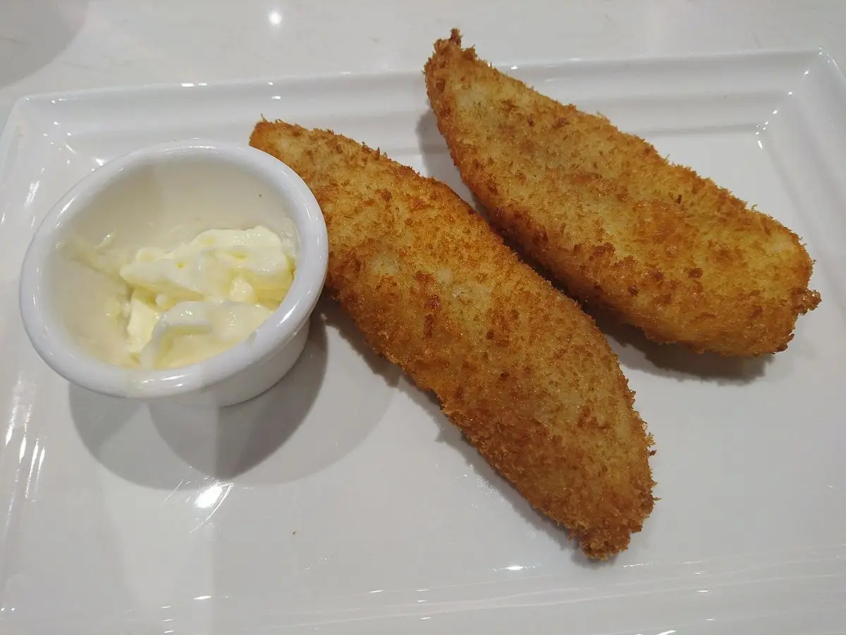 Dos filetes de pescado empanado dorado junto a un bol de salsa blanca sobre un plato