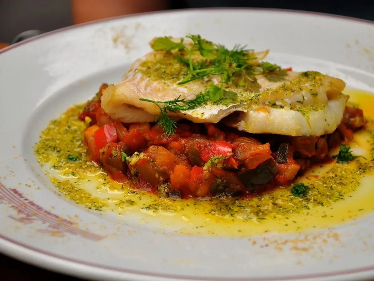 Lomo de bacalao a la plancha sobre un lecho de ratatouille de verduras con aceite de hierbas en un plato blanco