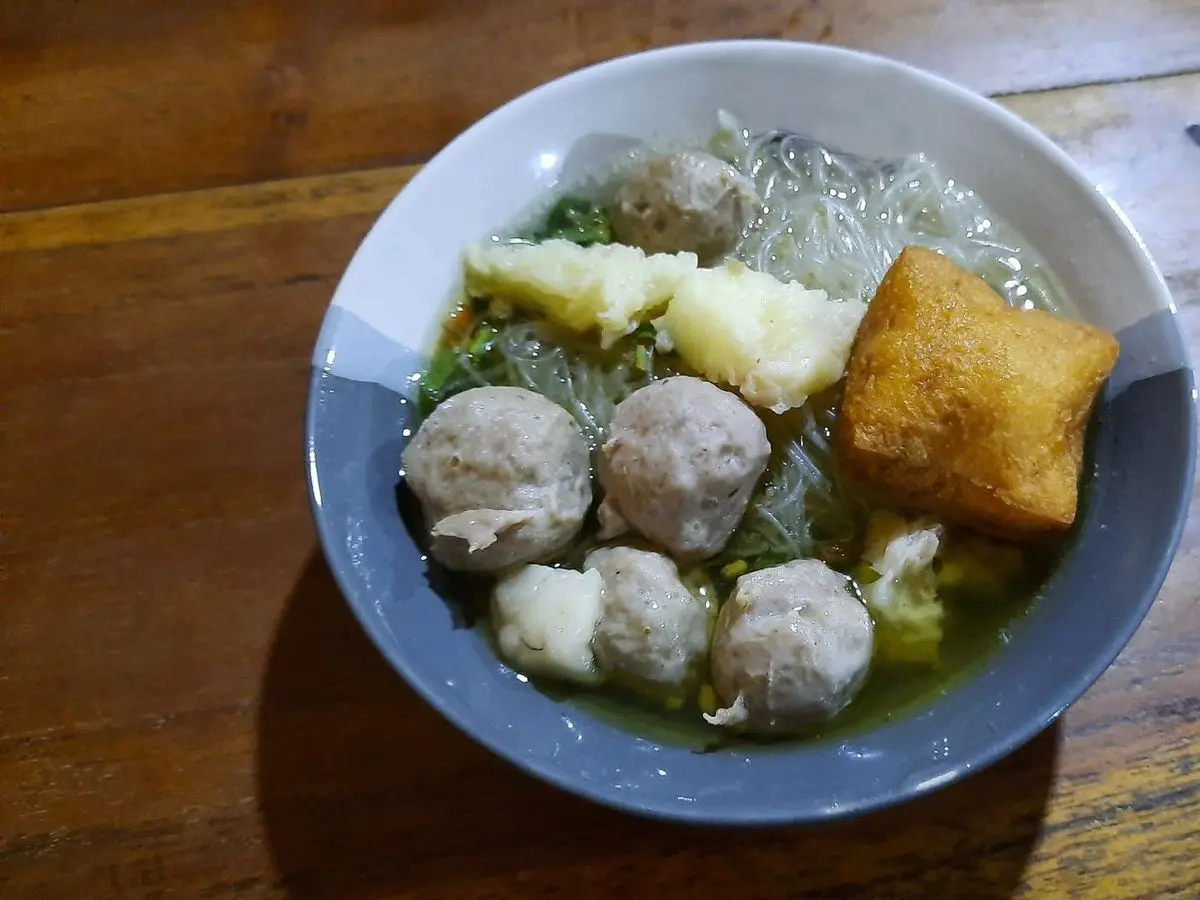 Cuenco de sopa de albóndigas con fideos transparentes, tofu frito y verdura sobre una mesa de madera