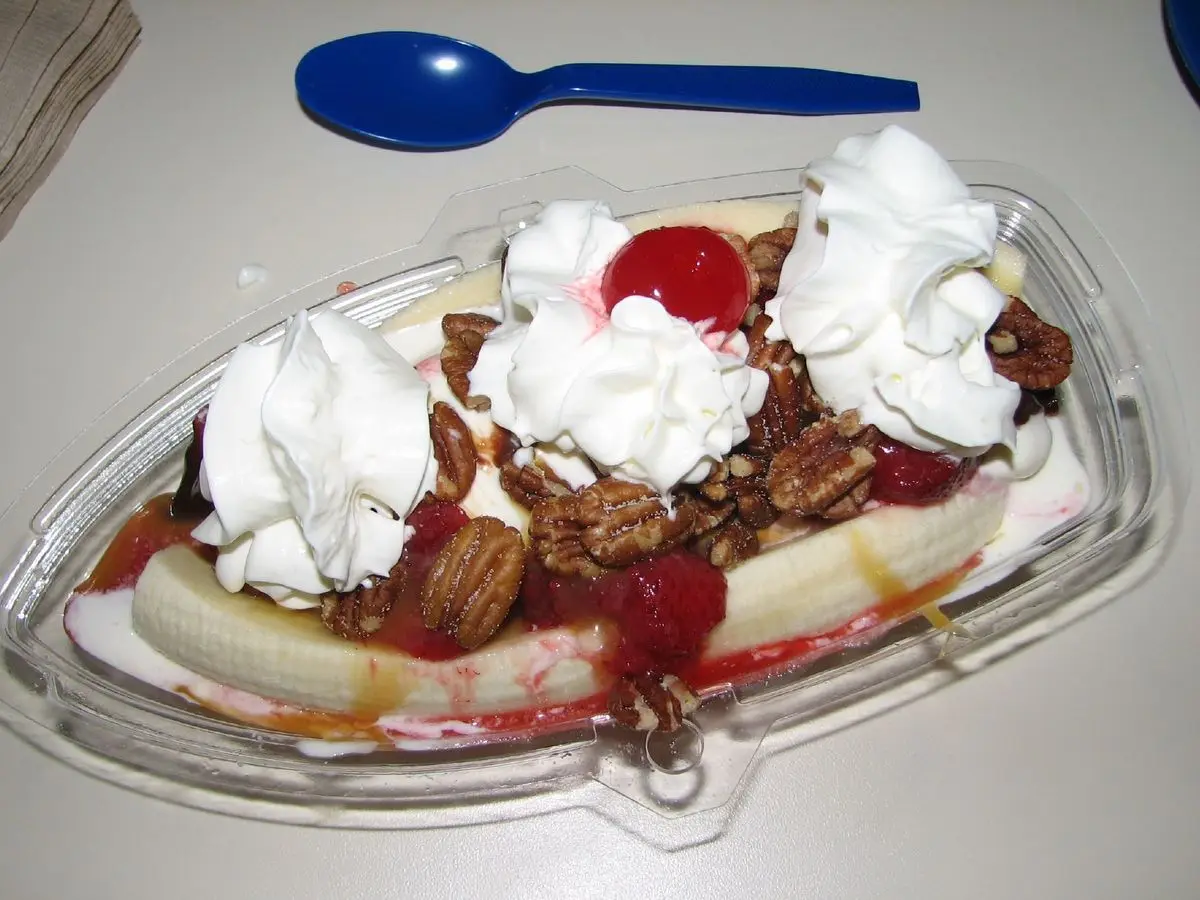Banana split en un bol alargado de plástico con nata montada, nueces y cereza