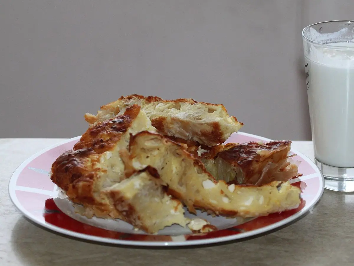 Porción de banitsa de hojaldre dorado en un plato junto a un vaso de yogur líquido