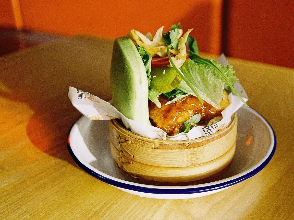 Bao relleno de pescado rebozado con lechuga, verduras y salsa en un cestillo de bambú
