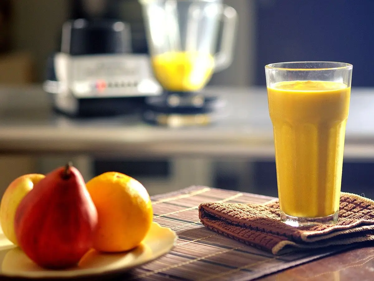 Vaso de batido naranja cremoso sobre una mesa con una pera y una naranja al fondo