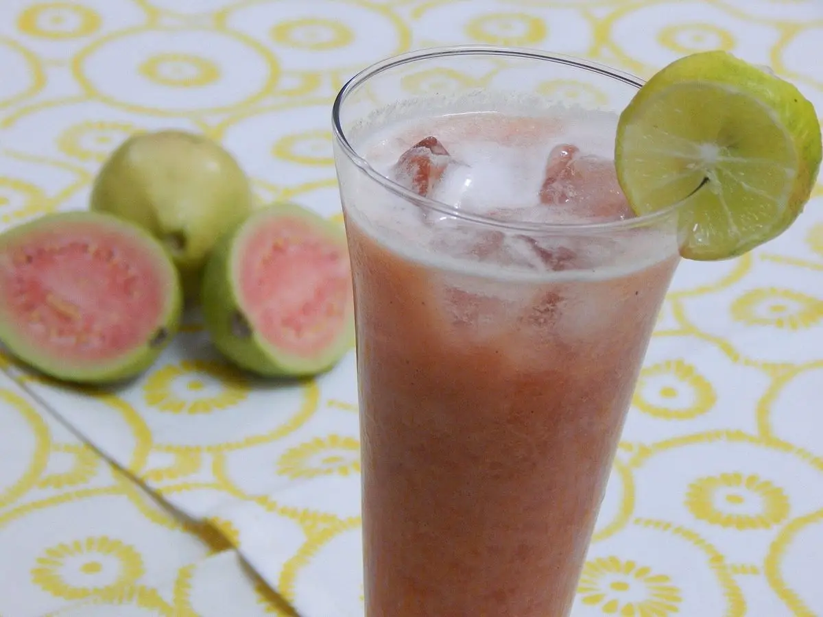 Vaso de bebida rosada de guayaba con hielo y una rodaja de lima, junto a guayabas abiertas