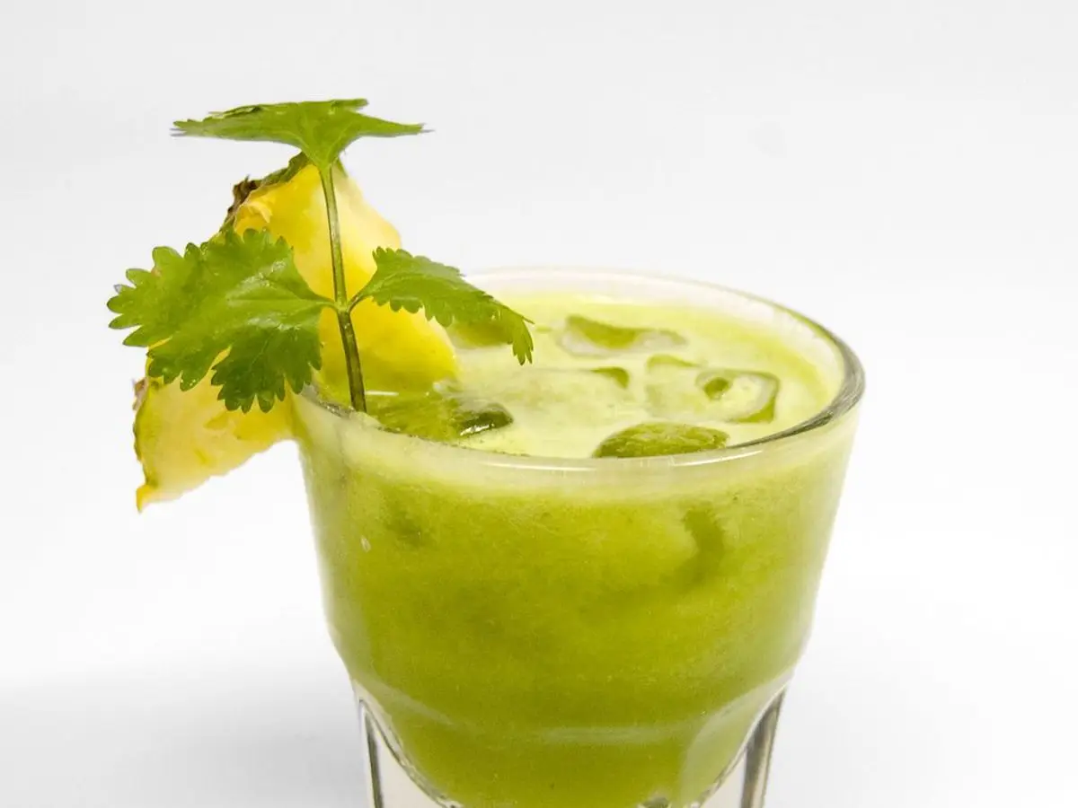 Vaso de batido verde con una rodaja de piña y hojas de hierbabuena sobre fondo blanco