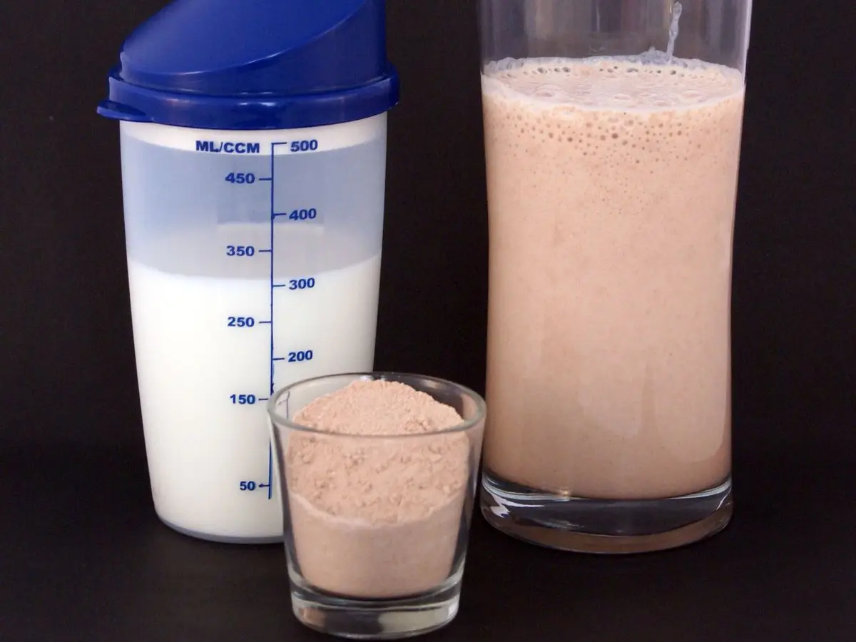 Vaso alto de batido proteico de color marrón claro junto a una coctelera azul y un cuenco con polvo