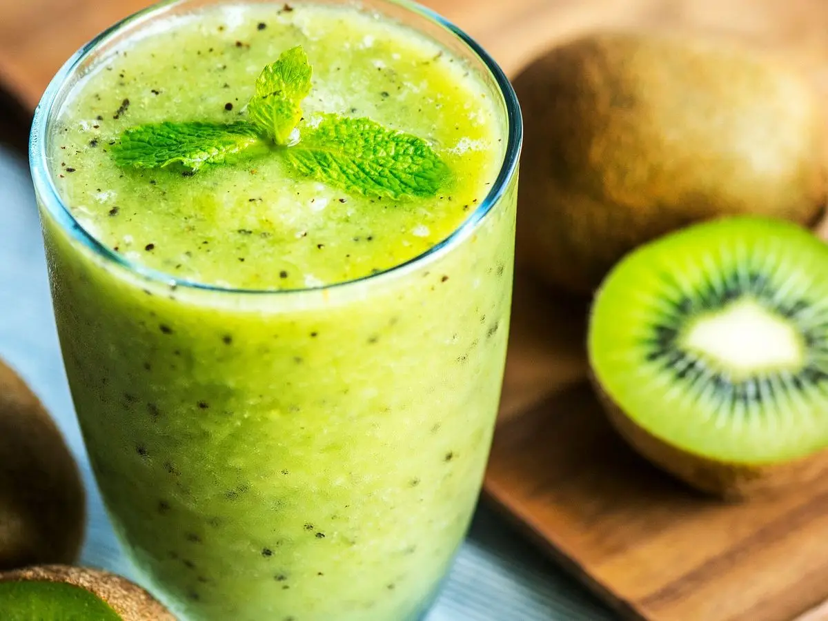 Vaso de batido verde de kiwi con hojas de menta, junto a kiwis cortados por la mitad