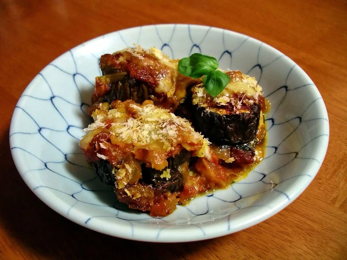 Plato blanco con berenjena asada gratinada con queso y tomate, decorado con una hoja de albahaca