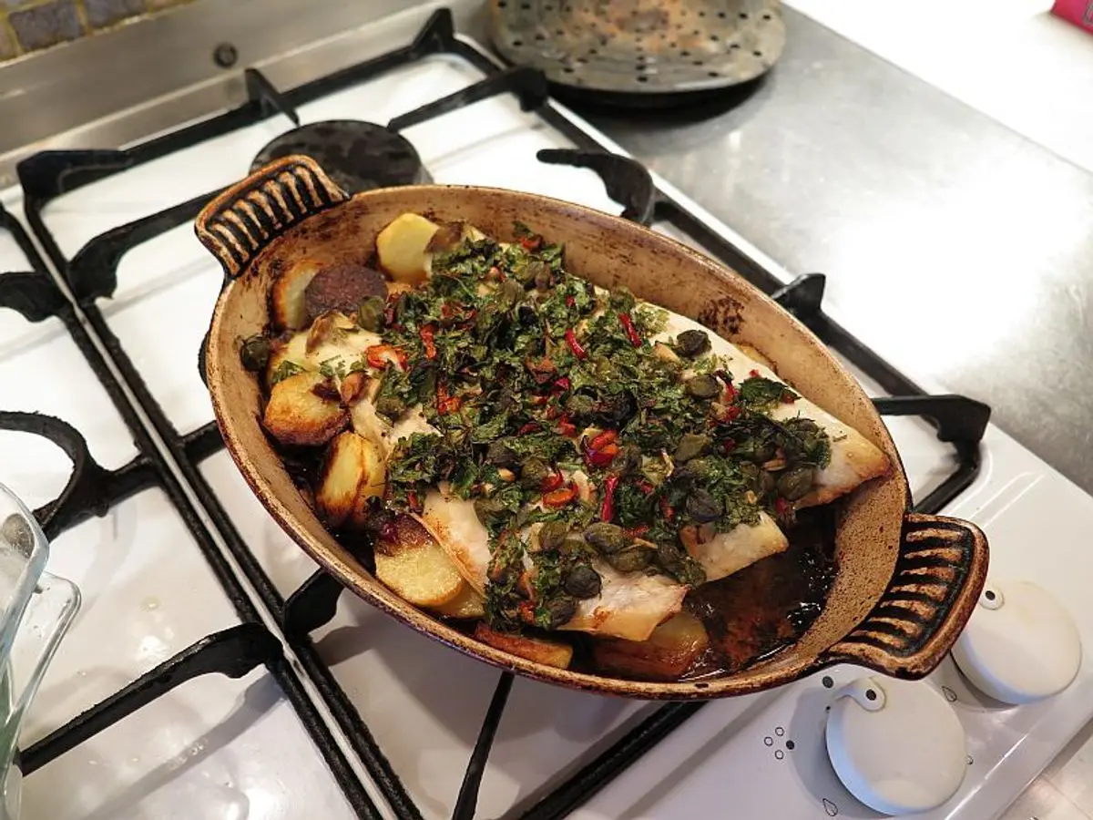 Fuente ovalada de pescado al horno con patatas, hierbas picadas y alcaparras sobre los fogones