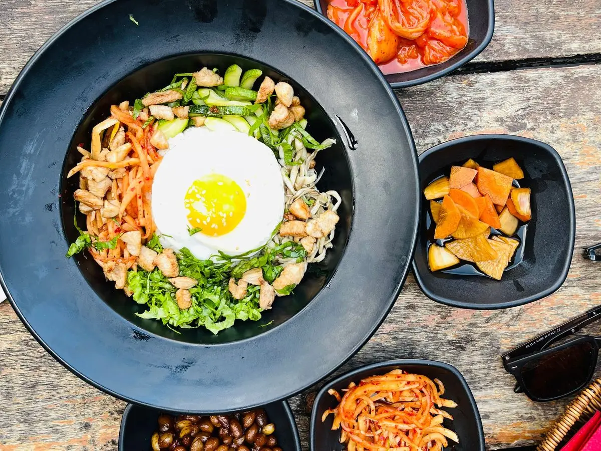 Bibimbap con huevo frito sobre arroz, zanahoria y verduras, rodeado de platillos de acompañamiento