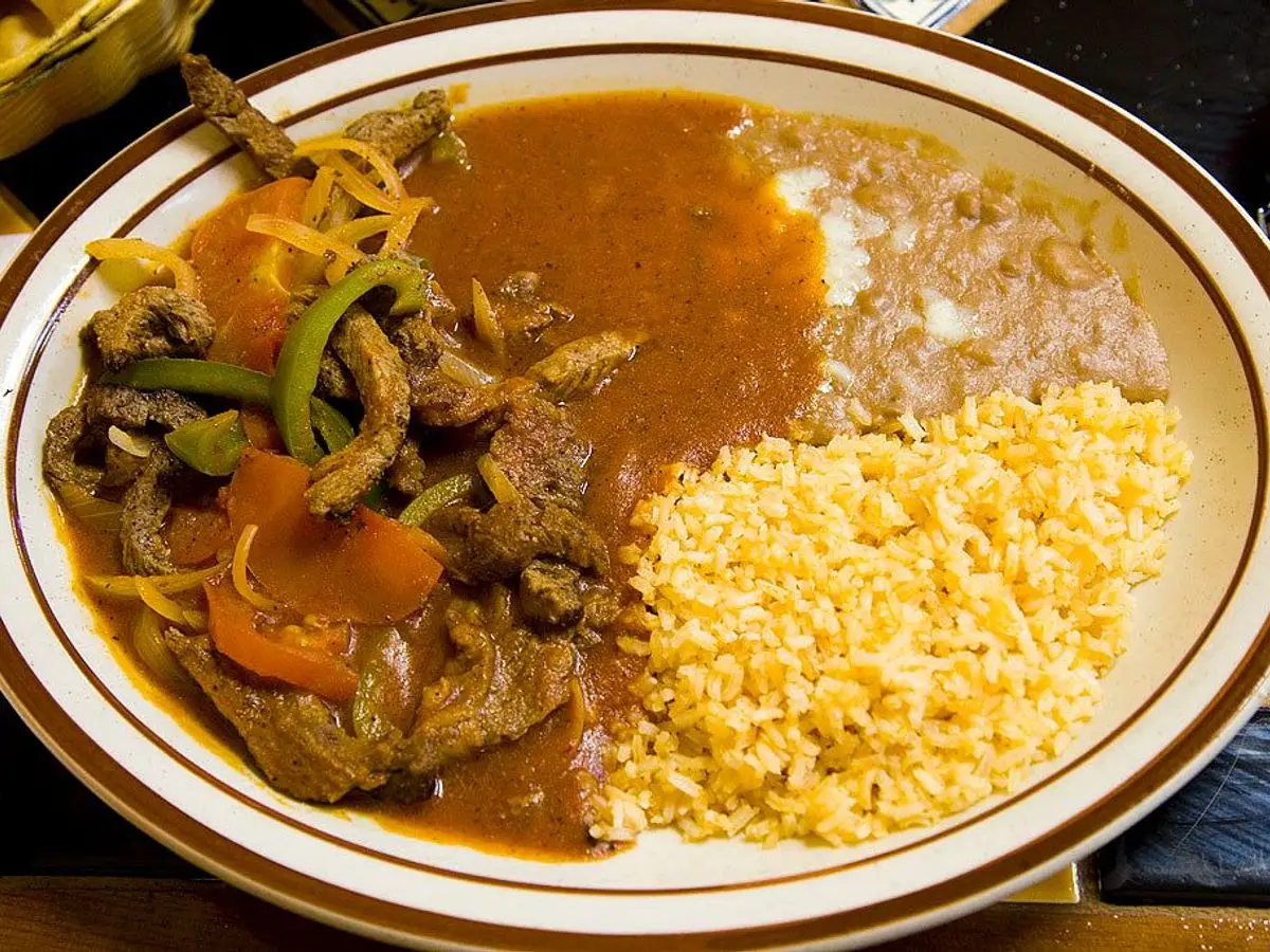 Plato con tiras de ternera guisadas con pimiento y tomate, frijoles refritos y arroz amarillo