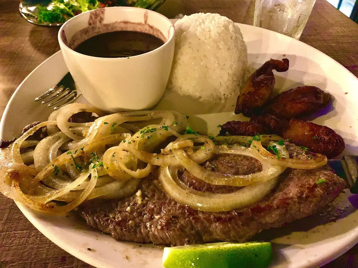 Filete de ternera cubierto de aros de cebolla con arroz blanco, frijoles negros, plátano frito y lima
