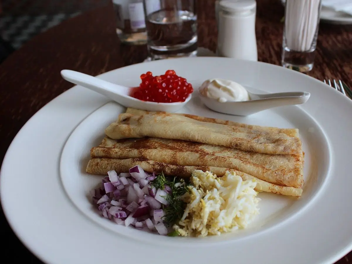 Blinis enrollados en un plato con cebolla morada picada, huevo duro rallado, crema agria y una cucharita de huevas rojas