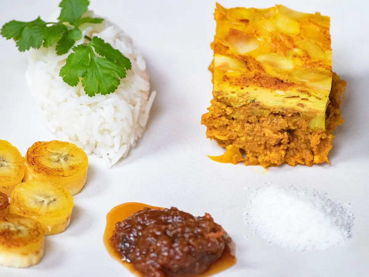 Porción de bobotie de carne especiada junto a arroz blanco, plátano frito y chutney