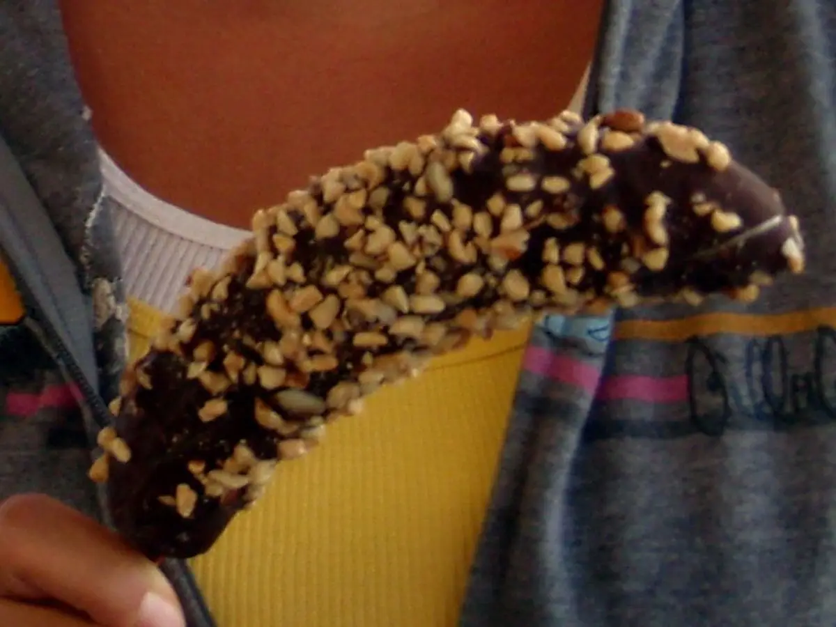 Plátano helado cubierto de chocolate y frutos secos troceados, sostenido en la mano