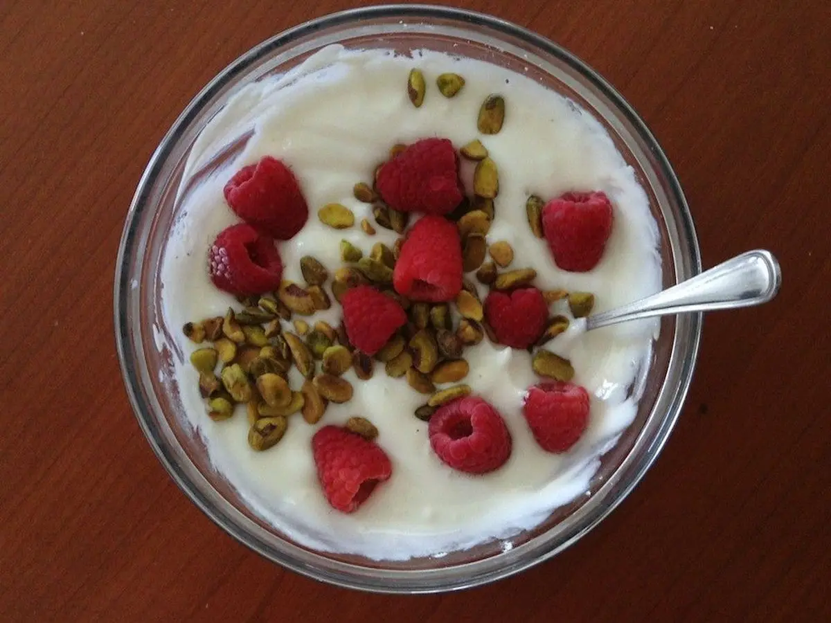 Cuenco de cristal con yogur, frambuesas y semillas por encima, con una cucharilla