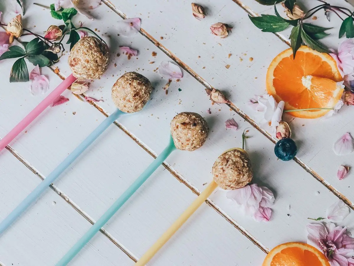 Bolitas de cereales y frutos secos pinchadas en palitos de colores sobre una mesa blanca con naranja y flores