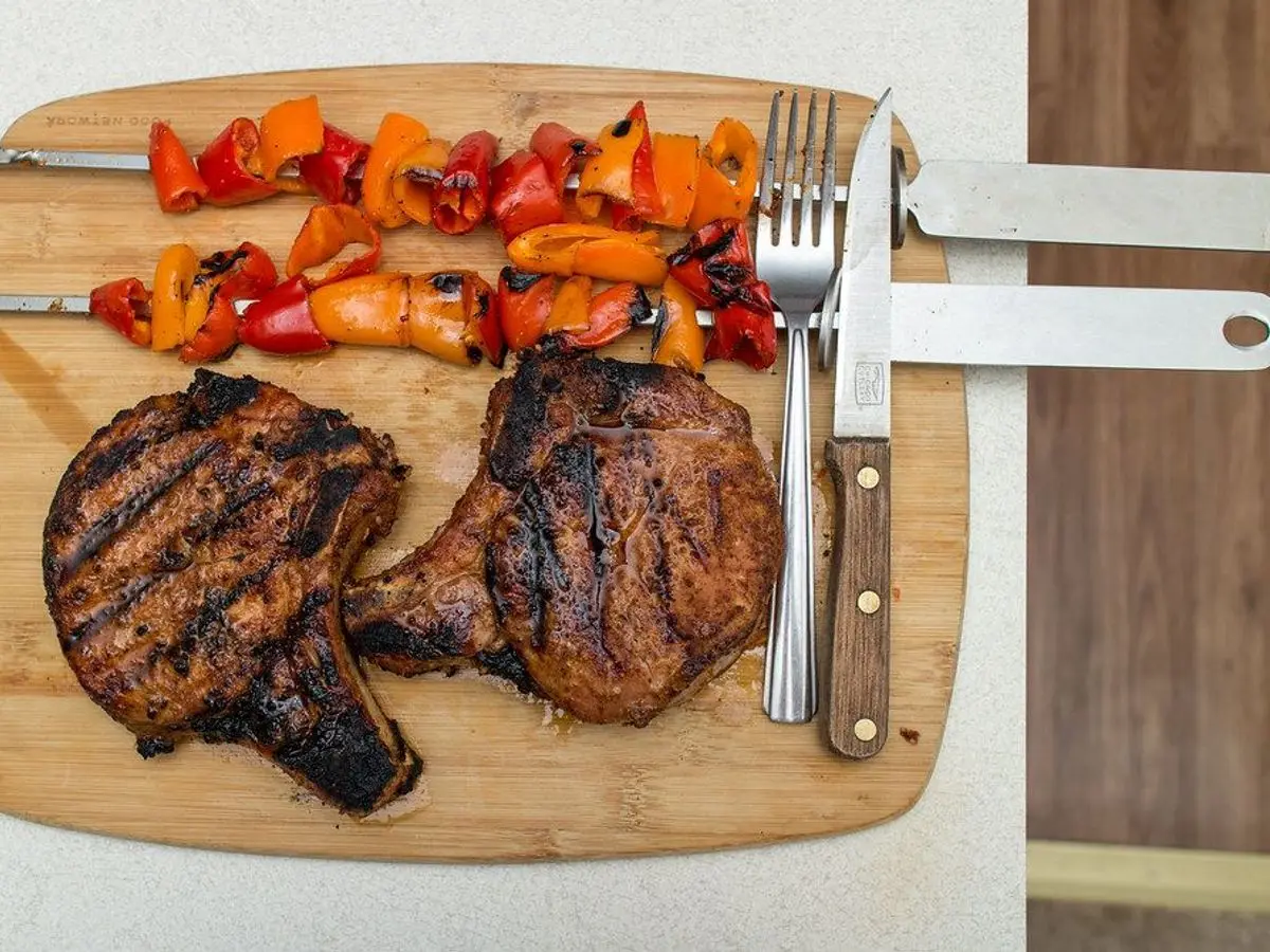 Chuletas de cerdo a la parrilla y brochetas de pimiento sobre una tabla de madera con cubiertos
